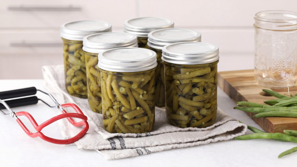 six canning jars filled with green beans cooling on a kitchen towel after being processed