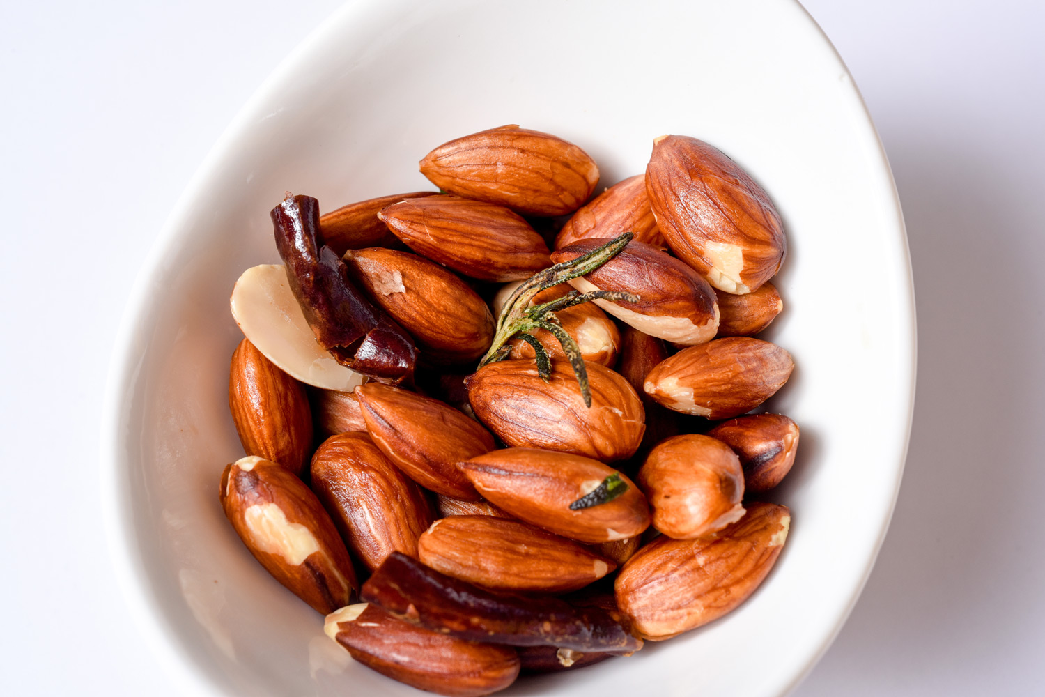 white bowl filled with roasted almonds, chili peppers, and a sprig of rosemary